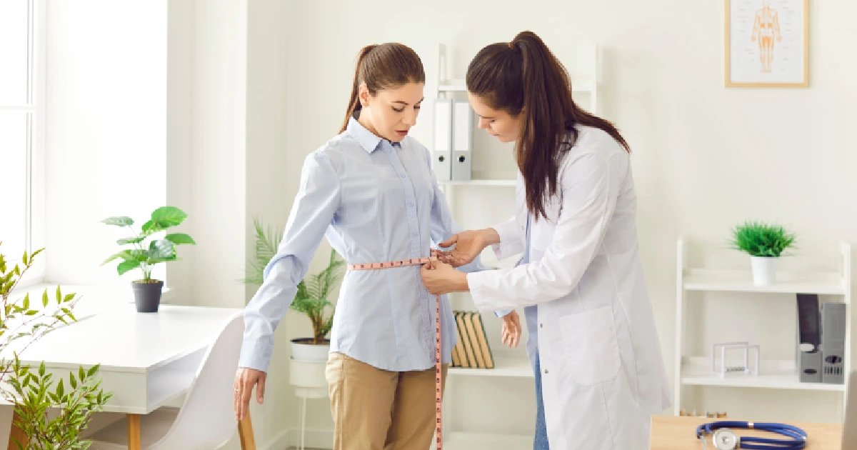 A healthcare professional measuring a woman's waist with a tape measure in a bright medical office in St Grand Blanc, MI, illustrating a medical weight loss consultation.